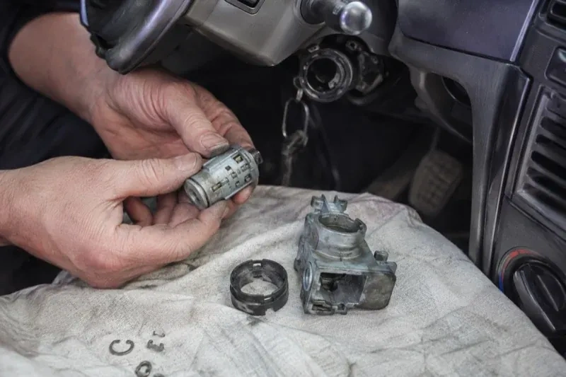 Hands holding a removed ignition cylinder beside a car steering column, with the housing and small parts on a cloth. — Ignition Lock Repair, Port Colborne